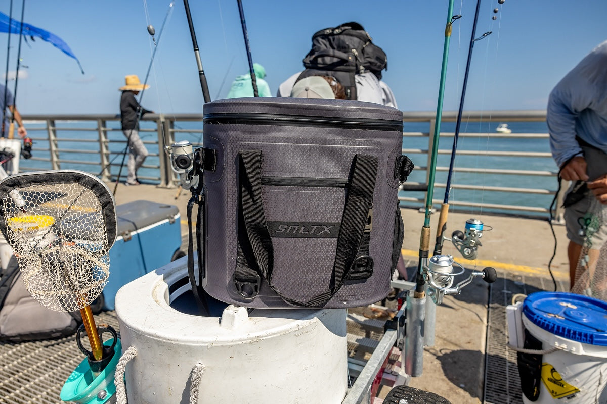 Gray cooler bag with 'Sultra' branding on a pier with fishing equipment and people in the background.