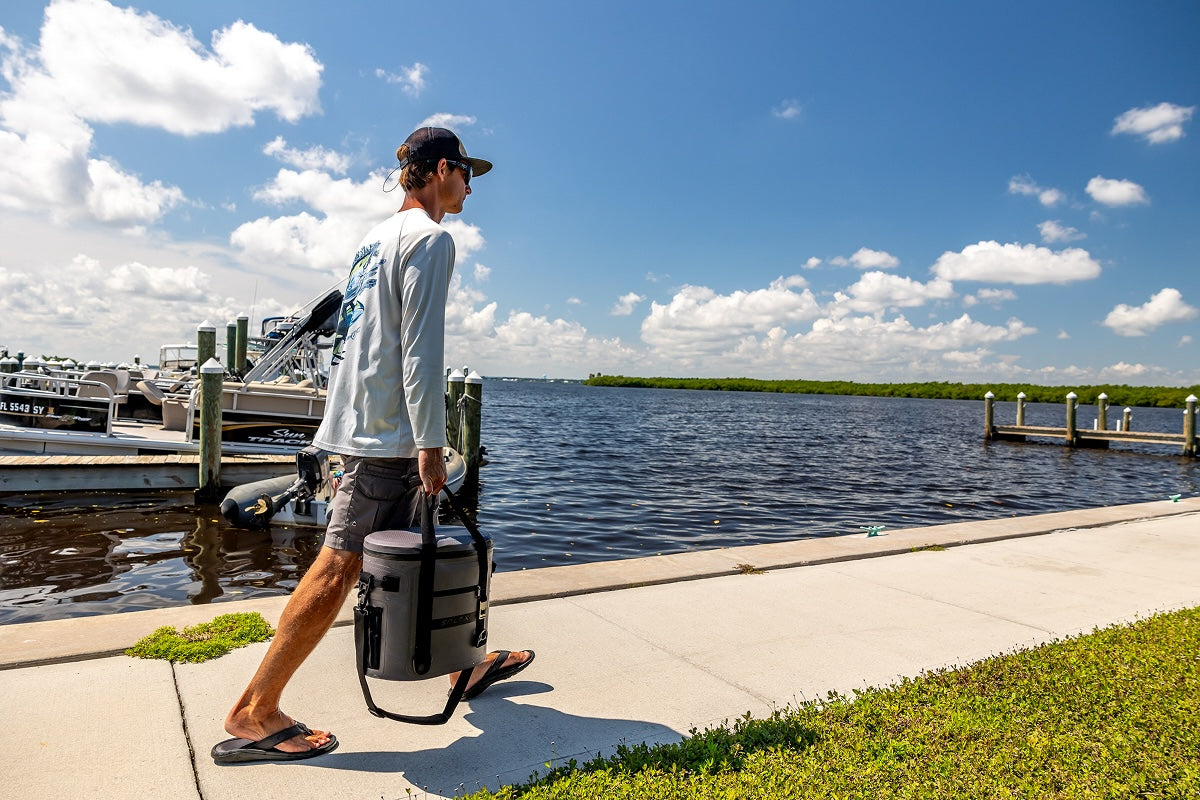 Man walking along a waterfront with a cooler bag, under a blue sky with clouds.