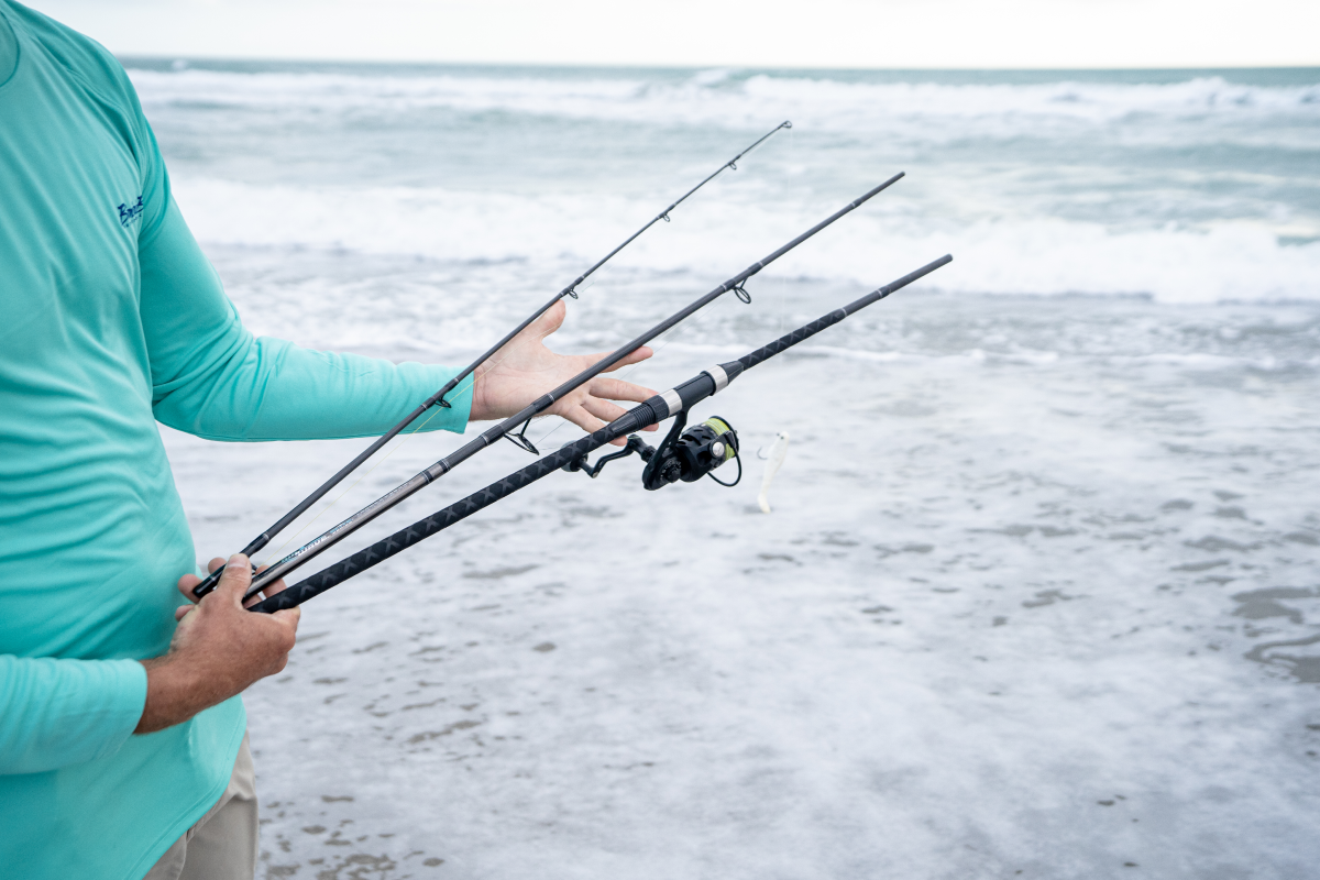 Person holding a three piece fishing rod on a beach with ocean waves in the background